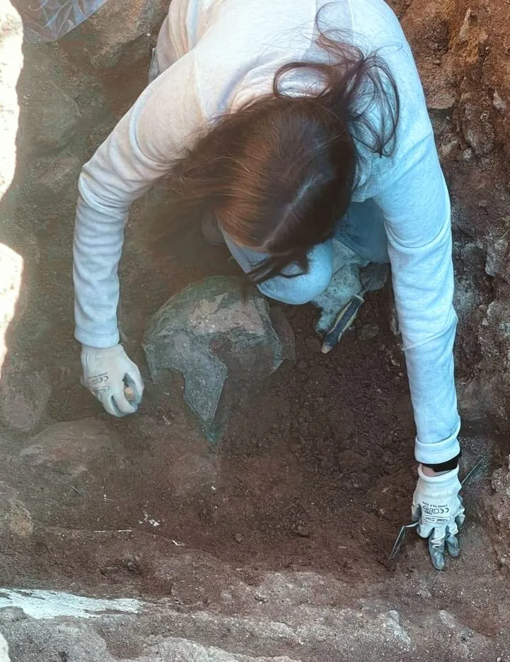 An archaeologist bends over the helmet, which is half-excavated and appears green due to oxidation