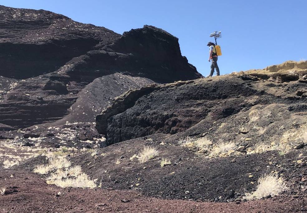 An image of a planetary scientist walking in a mountainous region with a yellow backpack on. The backpack has a camera attached to it.