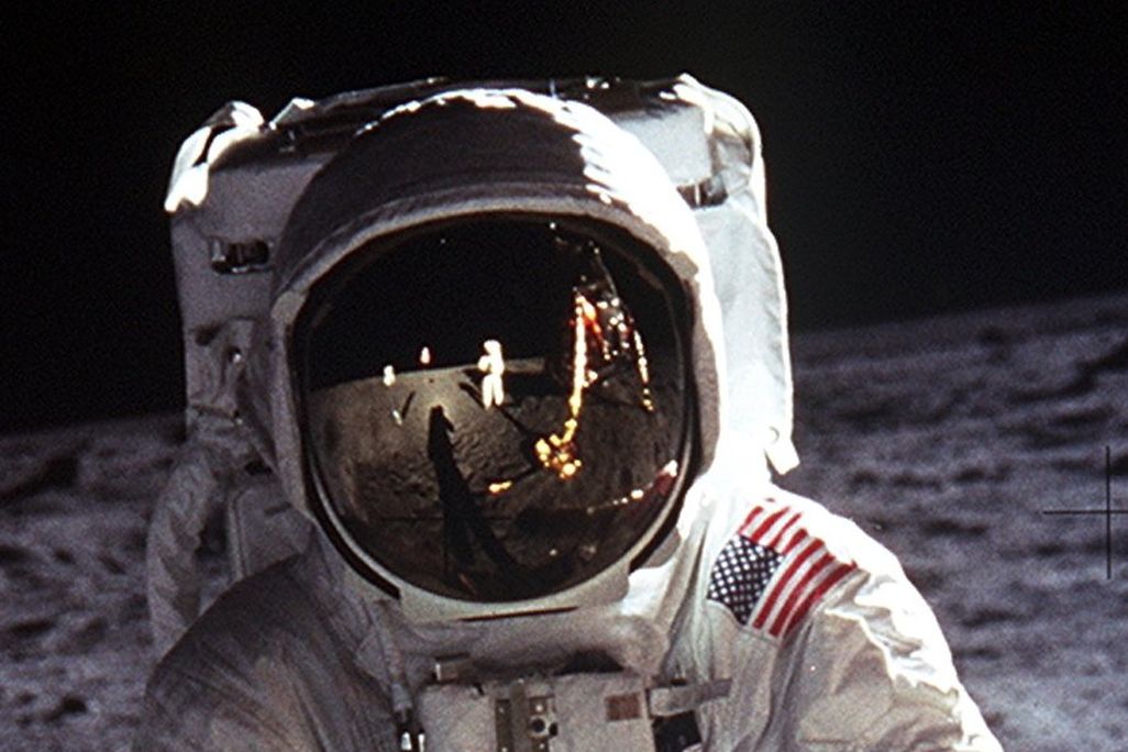 Astronaut Buzz Aldrin wears a large helmet and white space suit while standing on the moon. The American flag, Neil Armstrong and more of the moon is reflected in his helmet. The moon's rocky, gray surface makes up the background.