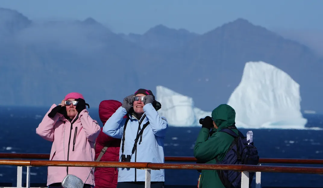 people wearing large jackets use eclipse glasses with an iceberg in the background