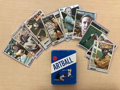 Baseball cards with faces of artists superimposed on them fanned out on a desk above a blue box with text that reads ARTBALL, an image of a batter, catcher, and umpire, with a red circle that says set 1.