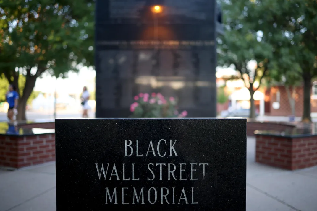 An outdoor memorial to the victims of the Massacre; foreground, a black slab with the words BLACK WALL STREET MEMORIAL engraved; in the background, a tall marble structure with names engraved on black stone