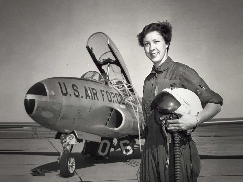 A photograph of a woman dressed to fly a jet, holding a helmet. The jet behind her has the top open and is labeled "U.S. Air Force"  