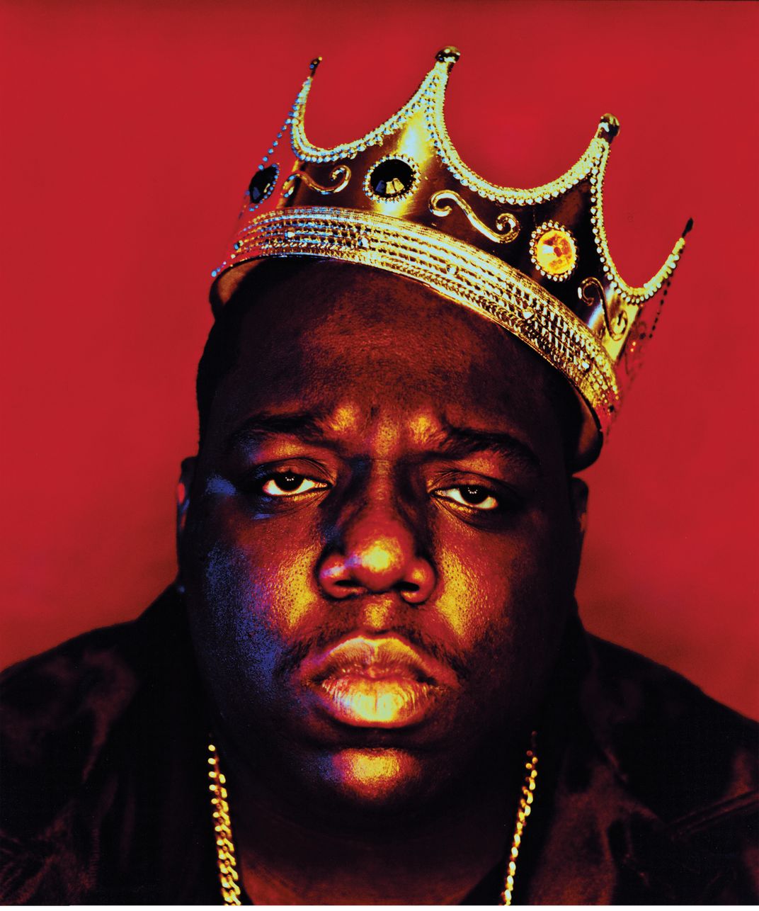 a Black man sits for a portrait wearing a gold crown in front of a red back drop
