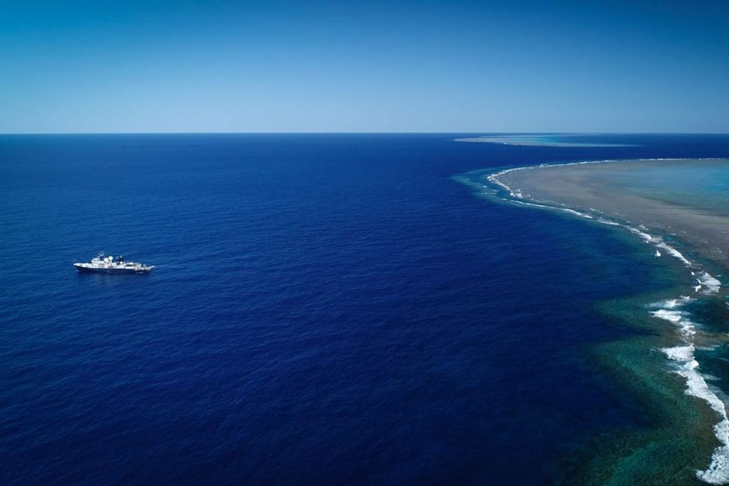 A photo taken from high above (likely by drone) shows the vast, bright blue ocean. The research vessel is on the left side. A long green and beige stretch of shallow coral reefs is on the right side. The horizon, where the deep blue ocean meets the lighte
