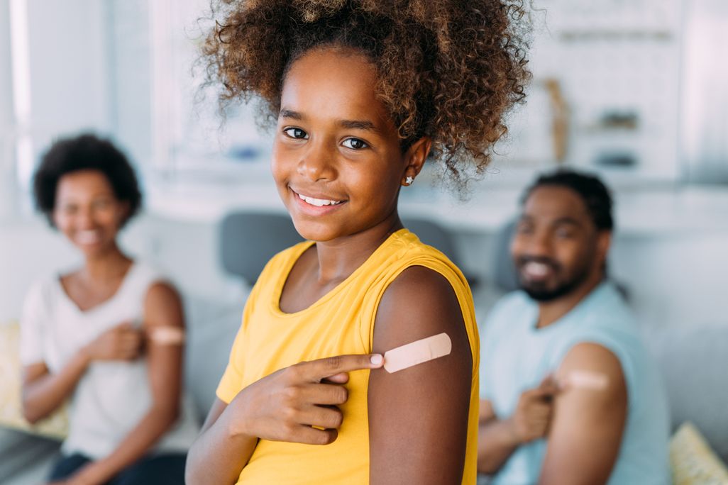 A young girl pointing to a bandaid on her left arm and smiling with parents in background