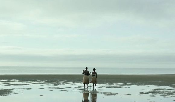 Two people in grass skirts stand on the shore, facing the ocean under an overcast sky.