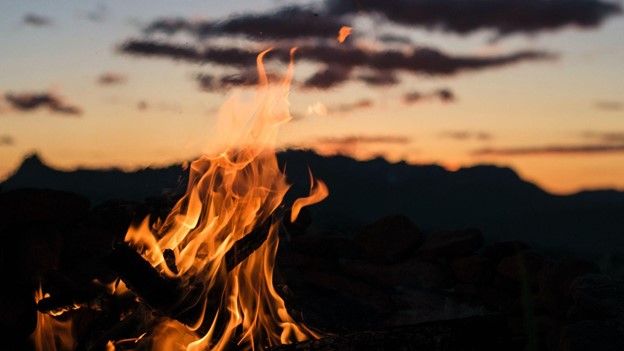 A small bonfire burns in the foreground with a sunset in the background.