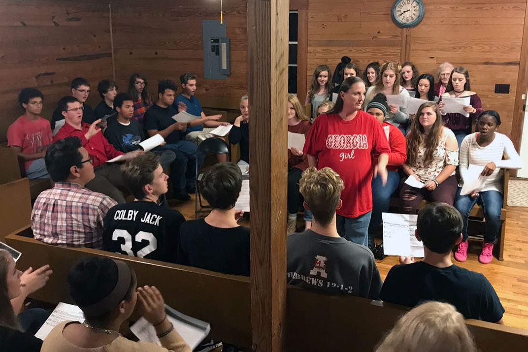 Students sit in pews on three sides of a woman in a red “Georgia Girl” shirt in the center.