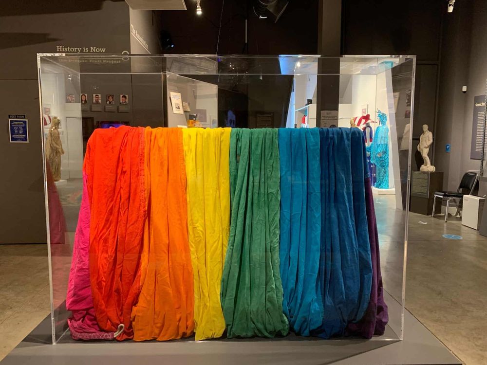 A view of a museum display case with the gathered folds of a piece of rainbow-striped cloth, including 8 bright colors from pink to purple