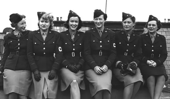 Six women sitting closely together posing for the camera. They are wearing uniforms with light skirts and dark jackets and an armband with the letter C on it.
