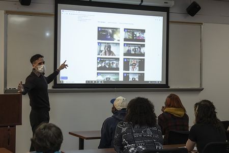 A man points to a video screen in a classroom with students seated in the foreground.
