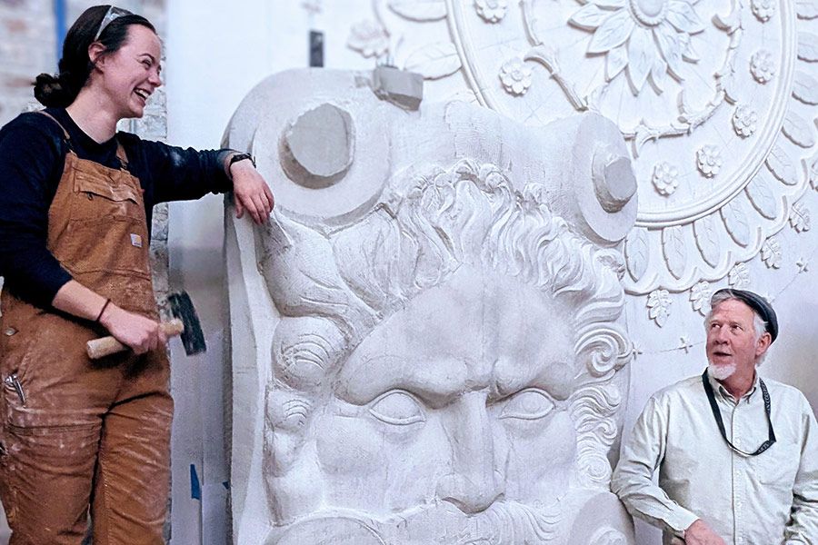 On either side of a large carving of a man’s face in white stone, a young woman holding a mallet and laughing and a man pose.