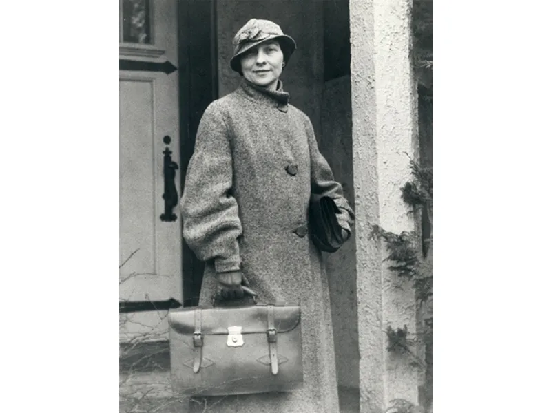 A young white woman in a long coat, wearing a fashionable hat cocked to one side on her head, carries a briefcase and poses in front of a doorway