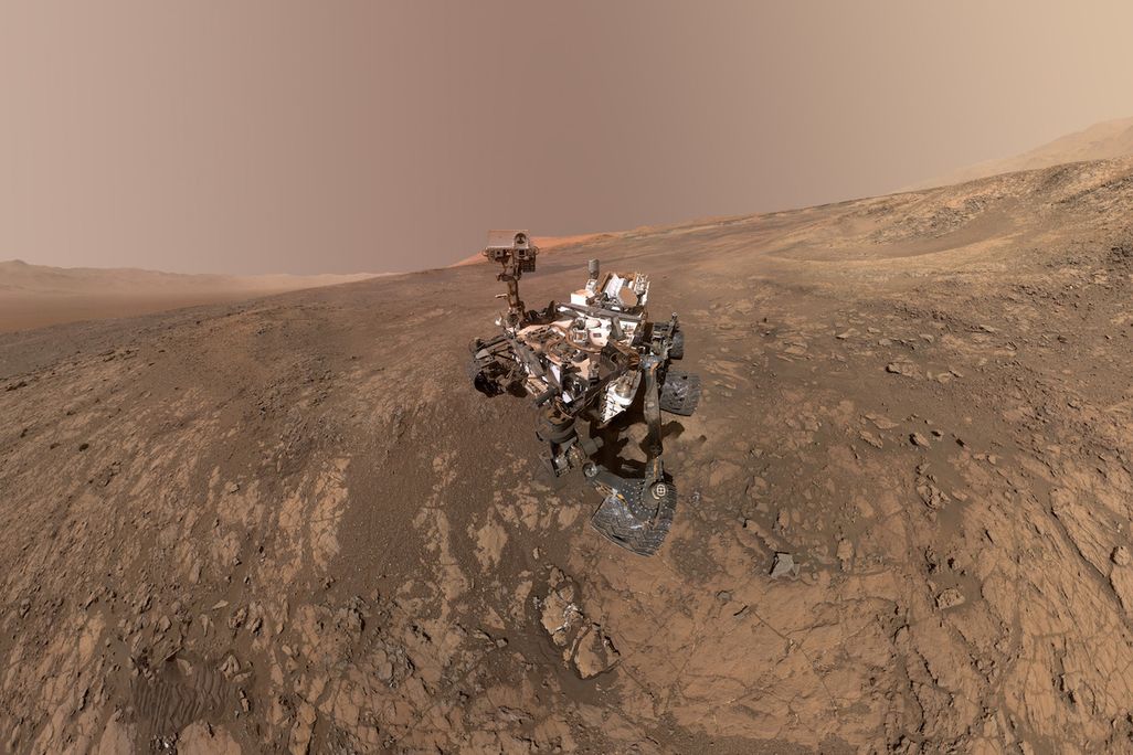An image of the Mars Curiosity rover on planet Mars with Mt. Sharp in the background. On the right, towards the horizon is the Gale Crater's rim.