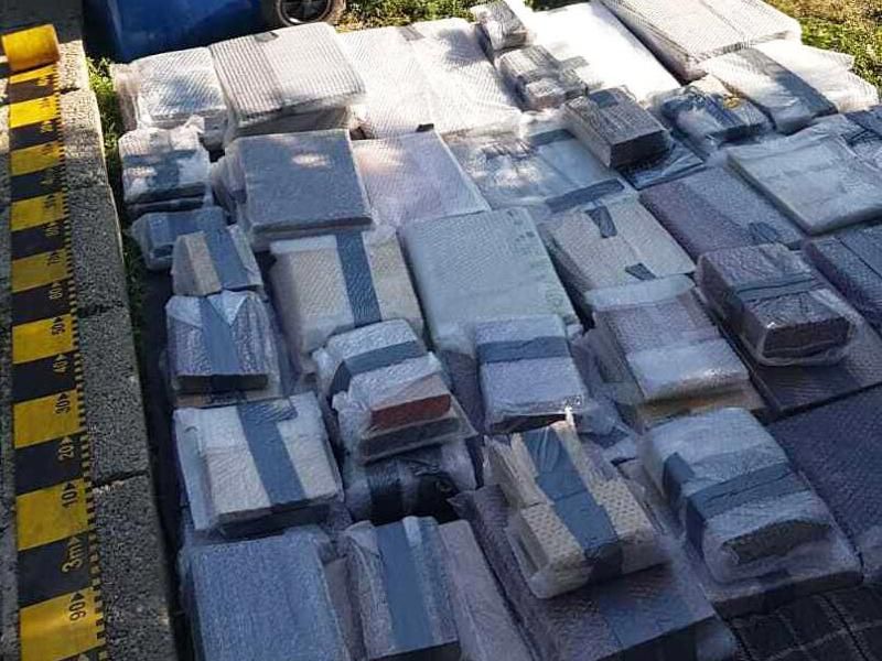 Stacks of books, which appear to be individually wrapped in protective fabric, lying on the ground in organized stacks next to a garbage bin and some plants