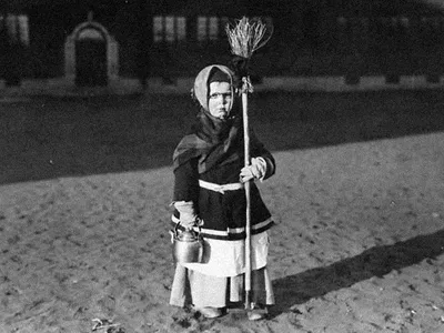 A young girl poses outside a building, wearing a dark coat, layers of skirts, and a kerchief over her hair, holding a broom in one hand and a kettle in the other. Black-and-white photo.