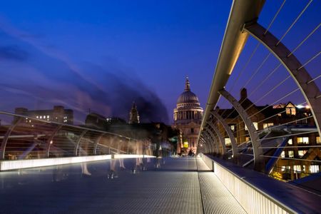 Pedestrians cross London's Millennium Bridge at dusk toward the lit dome of St. Paul's Cathedral.