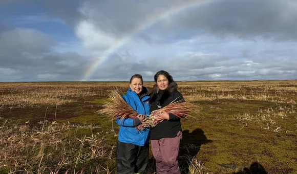 Two women stand in a field, holding bundles of grasses with a rainbow coloring the sky in the background.