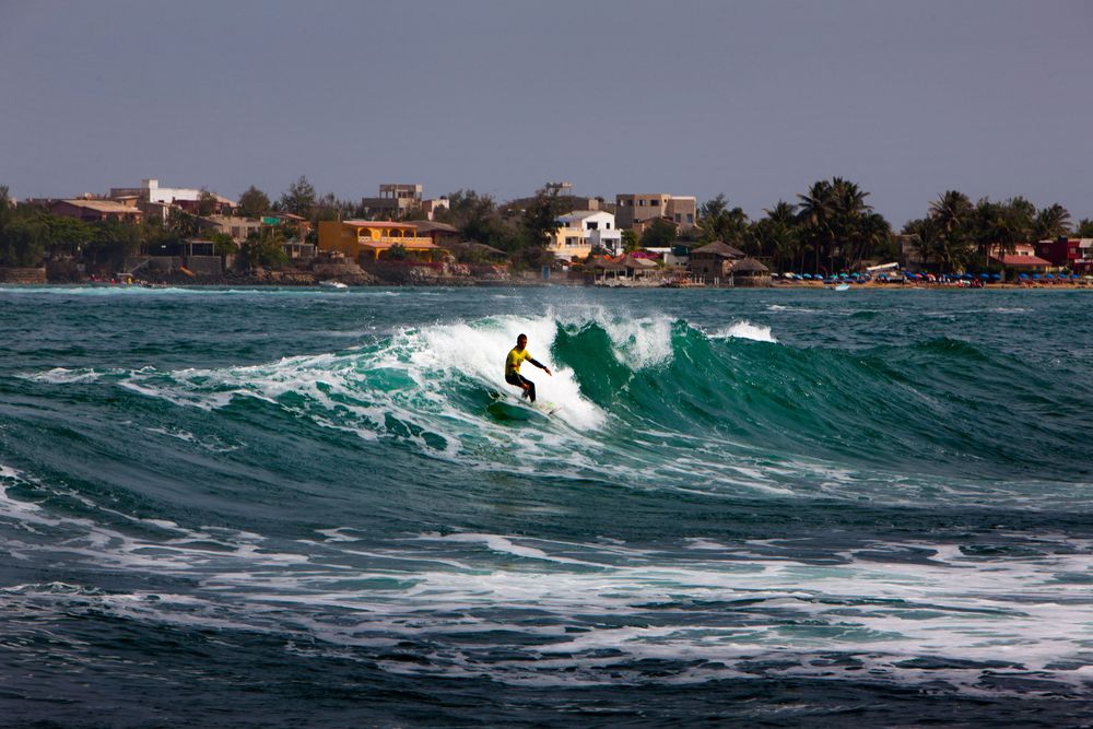 a surfer on a wave with some colorful buildings on the shoreline in the background