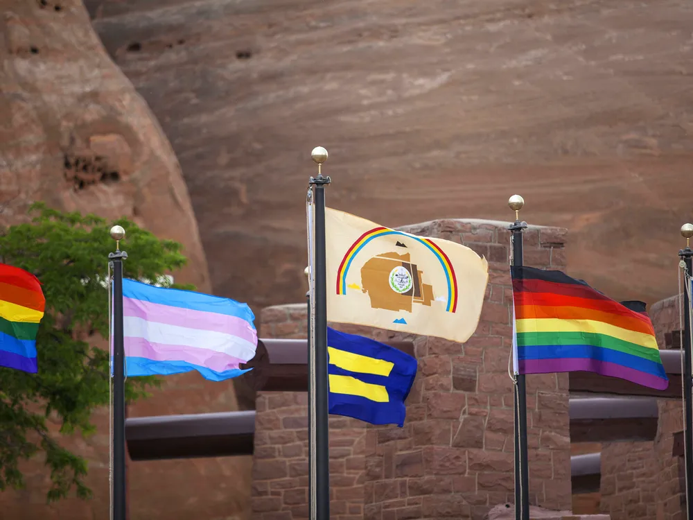 Various Pride flags (rainbow, transgender, equality, LGBTQ+ people of color, and love rainbow) displayed alongside the Navajo Nation flag in Window Rock, Arizona. (Photo by Pamela J. Peters (Diné)/Táchii’nii Photography, used with permission)