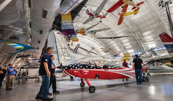 Inside a large hangar filled with natural light, three white people maneuver a small red, white, and blue airplane into its display space. At least eight aircraft are suspended from the hangar's ceiling.