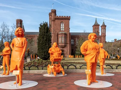 Five orange, 3D-printed statues of female scientists in a garden in front of the Smithsonian Castle on a sunny day.