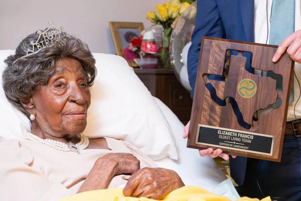Elizabeth Francis, a Black woman, in bed with a "happy birthday" tiara and a plaque that reads "Elizabeth Francis, oldest living Texan"