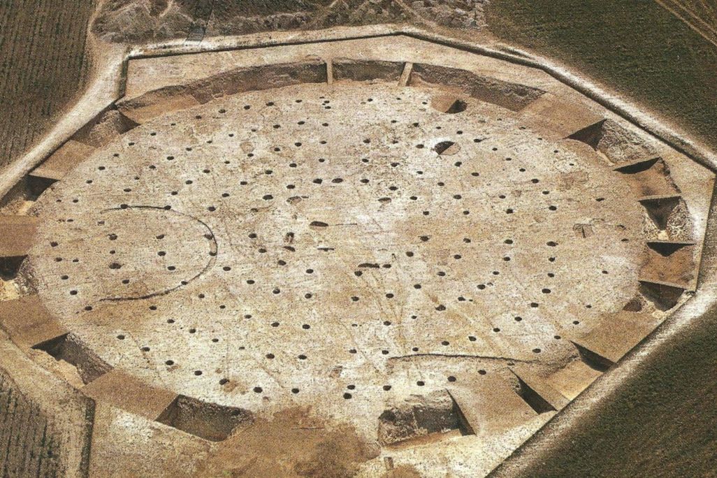 An aerial view of sandy dirt ground with numerous holes in concentric circles, and a ditch surrounding the entire formation
