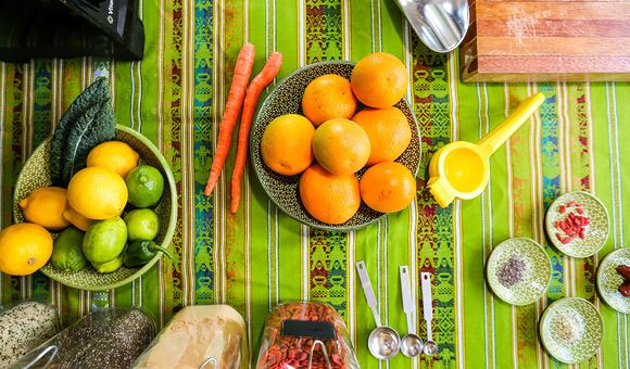 Bowls of citrus fruits including lemons, limes, and oranges, are arranged on a striped, bright green table cloth. Behind the bowls are jars filled with various superfoods including Goji berries.