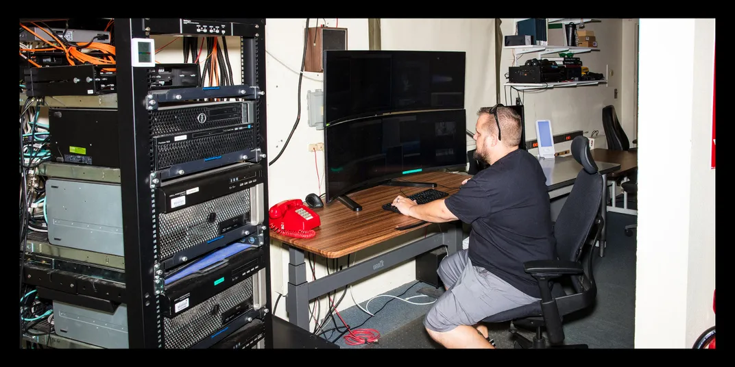 Rankin sits at a desk with a wide, curved computer screen and a metal shelf of hardware