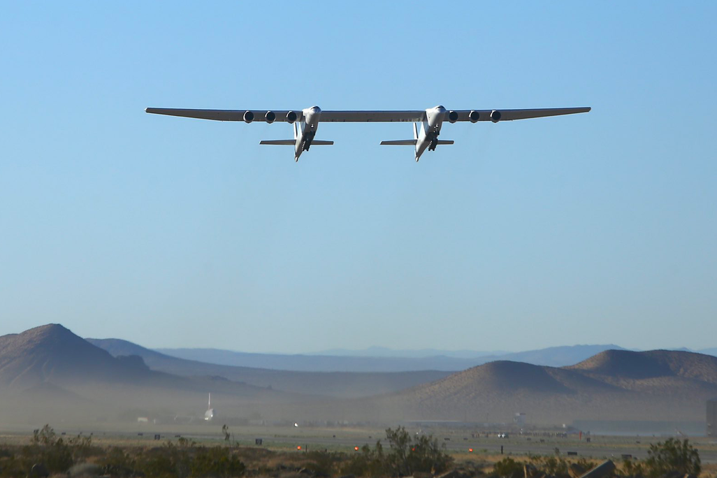 The 385-foot-wide Roc aircraft flies in a clear blue sky above the desert with mountains on the horizon