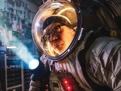 Adam Savage poses heroically in his spacesuit, which appears white, bulbous, and dusty despite never going to space. His face, wearing glasses, is illuminated behind the transparent plastic bubble that forms most of the helmet.