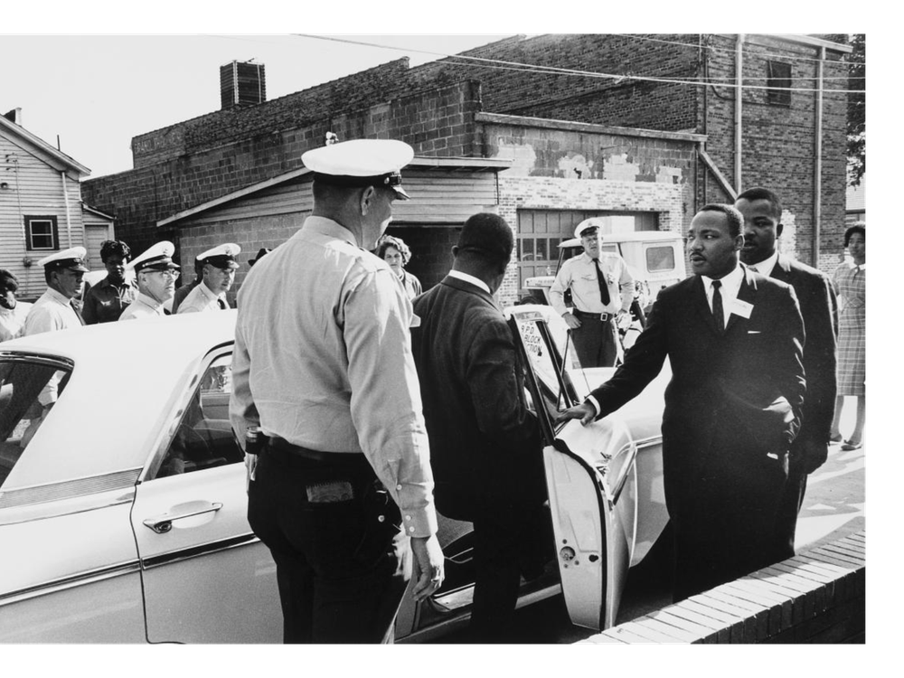 A black and white photograph of policeman in foreground, one man stepping into a squad car and MLK with an arm outstretched moving toward the car