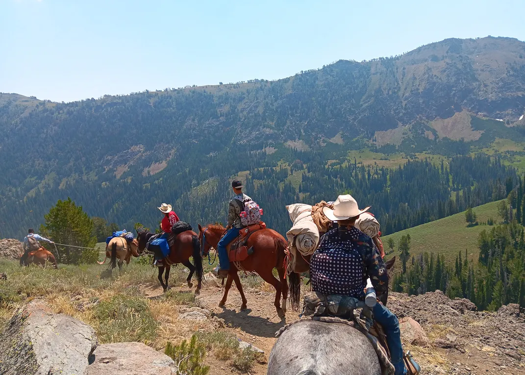 Four people ride on horseback carrying lots of gear through green mountains.