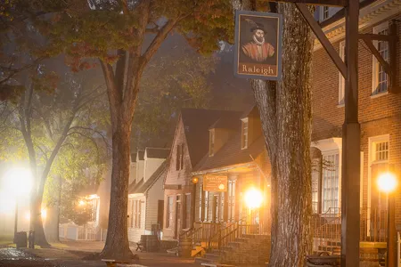 A foggy autumn evening in Colonial Williamsburg, where glowing storefronts and historic architecture create a warm and timeless streetscape.&nbsp;