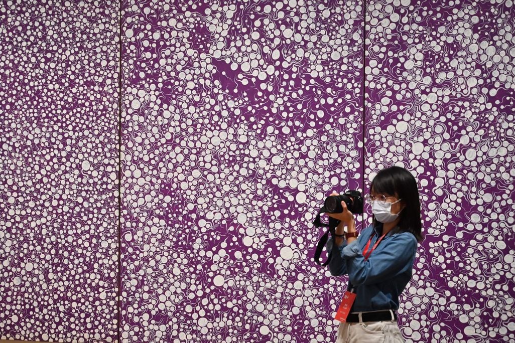 Woman wearing a mask and taking a photo in front of a purple background with white polka dots