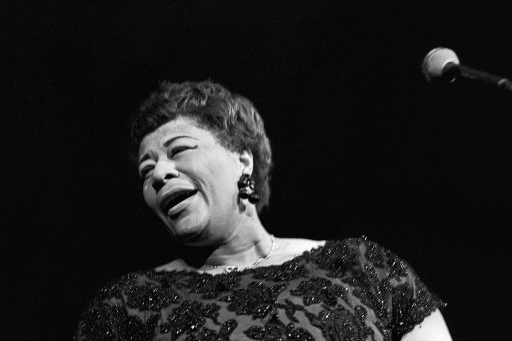 A black and white image, closely cropped to Fitzgerald's profile with the microphone just peeking in on the side; Fitzgerald is singing and turns away from the camera, smiling, mouth half-open and eyes closed, wearing a beaded dress and short cropped hair