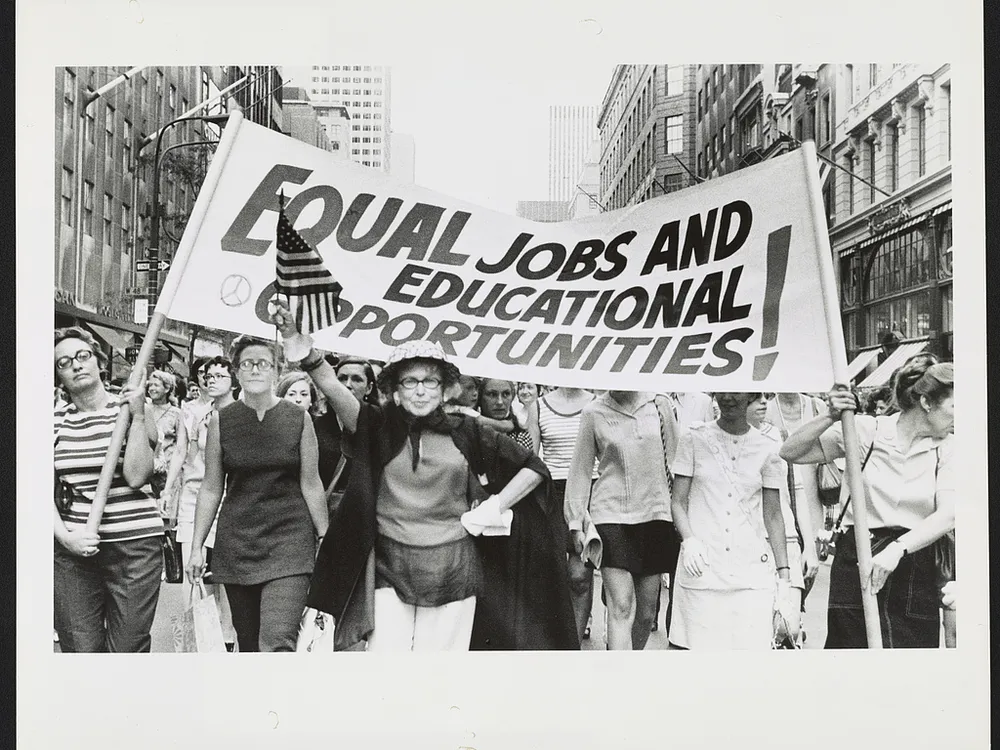 Women marching down the street holding a banner with the message “Equal jobs and educational opportunities!”