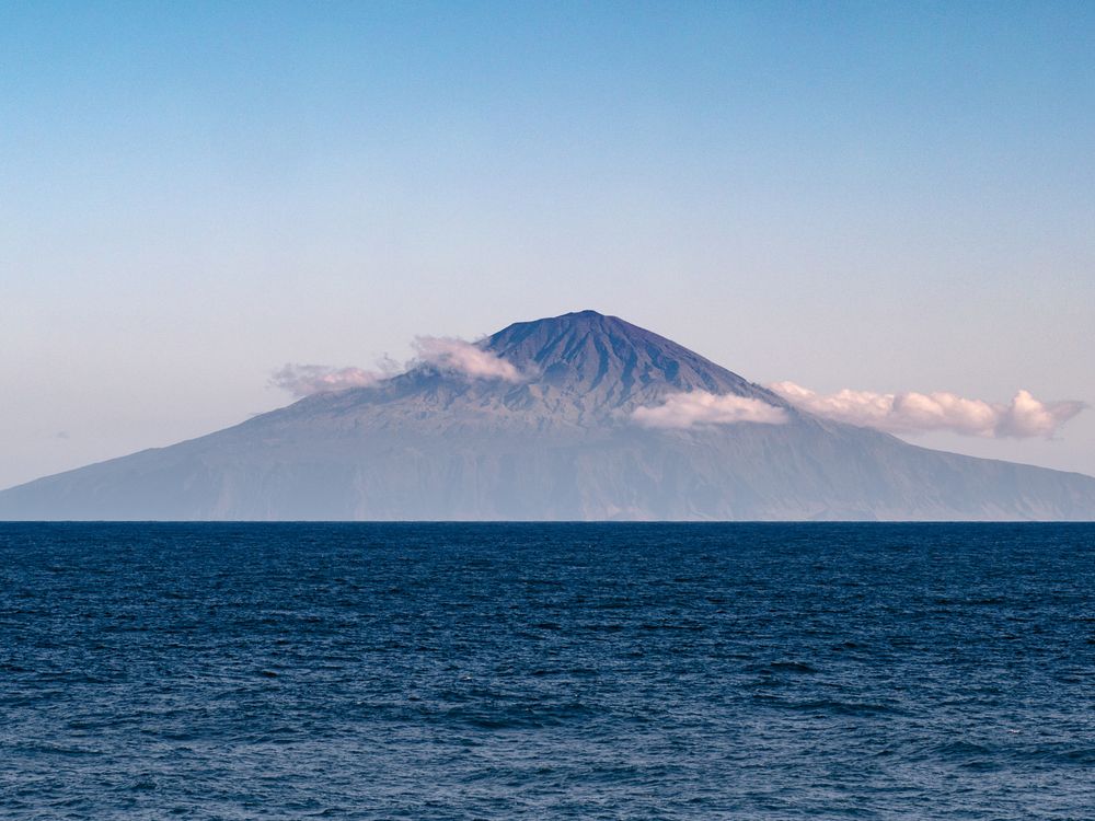 A photograph of Tristan da Cunha taken from a boat out at sea. The blue ocean water meets the horizon halfway up the photo. The island, shaped like a flat triangle sits at the horizon. It has small clouds in front of it and a blue-pink sky in the backgrou