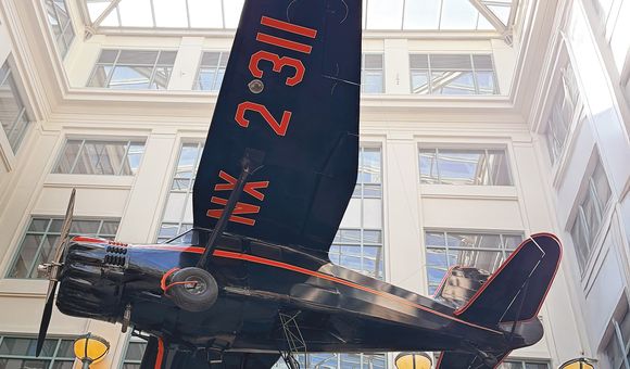 An upward-looking shot shows a dark-colored, single-engine Stinson Reliant monoplane with red markings on its wing, suspended indoors from the ceiling of a multi-story building with a glass atrium roof.