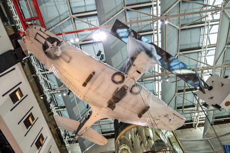 A World War II military airplane is photographed from below while hanging from the ceiling. The underside of the airplane is painted white and the wing and forward fuselage are wrapped in opaque plastic.