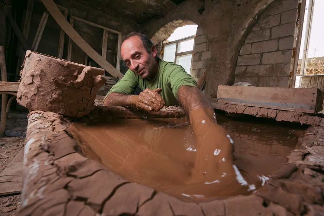 A man in a green shirt reaches in to a pool of clay, prepared for work.