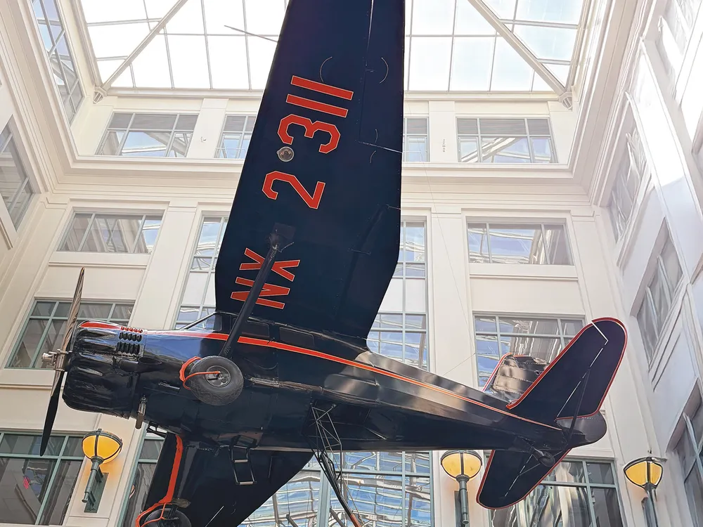 An upward-looking shot shows a dark-colored, single-engine Stinson Reliant monoplane with red markings on its wing, suspended indoors from the ceiling of a multi-story building with a glass atrium roof.