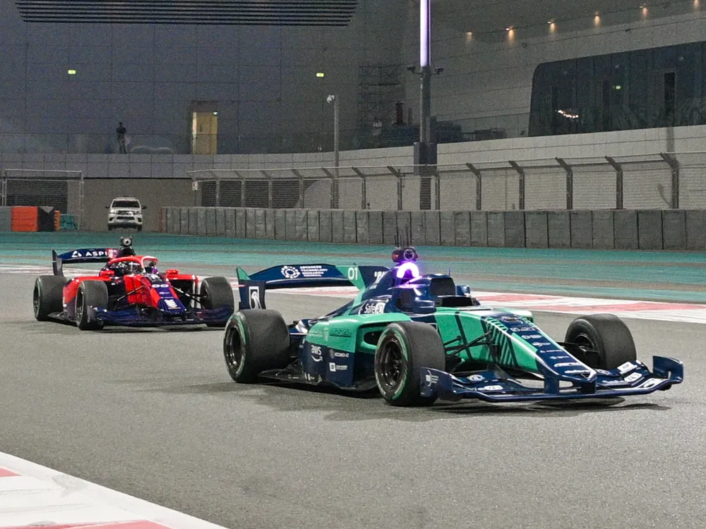 Two driverless race cars, one green (foreground) and one red (background) speed down a straight on the Yas Marina circuit.