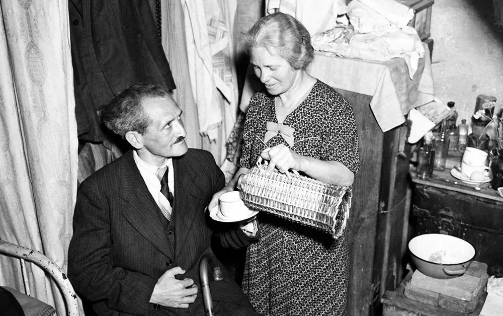 A white woman in a dress pours tea for a seated man in a suit, in a small home crowded with items (longform main)