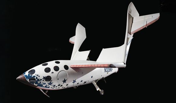 A white spacecraft against a black background. The fuselage is spangled with dark blue stars and black circles. A large wing is swiveled up away from the fuselage. The tail of the spacecraft is marked by this identifying number: N328KF.
