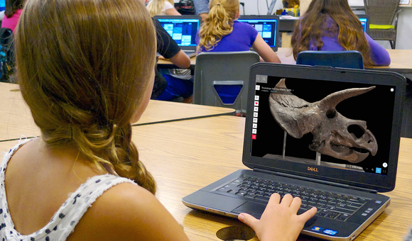 Girl in a classroom using a laptop computer that shows a dinosaur skull on its screen. Several other students work at desks with laptops in the background.