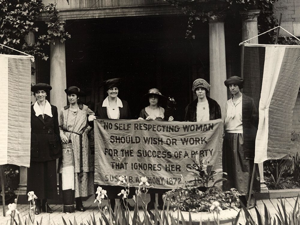 Suffragists stand holding a banner quoting Susan B. Anthony: "No self respecting woman should wish or work for the success of a party that ignores her sex."