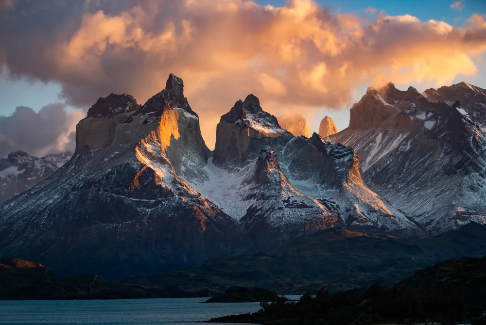 A dramatic, color landscape of Torres del Paine at sunrise. The iconic granite peaks are bathed in intense, fiery light under cloud cover, emphasizing the grandeur of the Patagonian mountains rising above the dark lake.